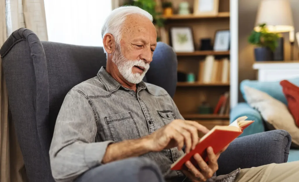 A senior man sits in a chair reading a book.